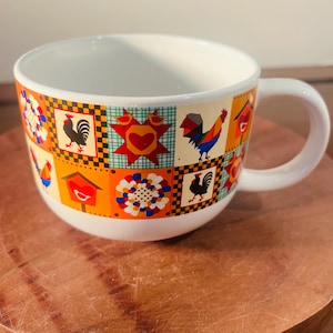 Puede incluir: Una taza de sopa de cerámica blanca con una banda estampada colorida con gallos, casas para pájaros y diseños florales. La taza tiene un asa grande y está colocada sobre una superficie de madera. El diseño incluye cuadrados naranjas, amarillos, rojos y negros.