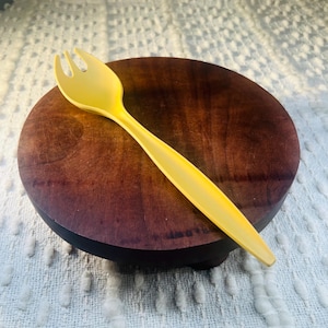 Puede incluir: Un tenedor de plástico amarillo con tres púas descansa sobre una tabla de servir de madera marrón oscuro. El tenedor tiene un mango largo y delgado. La tabla de servir es redonda y se asienta sobre una pequeña base negra. El fondo es una superficie blanca texturizada.