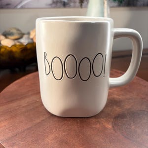 Op de afbeelding: Een crèmekleurige keramische mok met een afgeronde, licht onregelmatige vorm en een stevig handvat. Het woord BOOOOO! staat in zwarte, dunne, handgeschreven letters op de voorzijde van de mok. Het oppervlak heeft een gladde, matte afwerking.