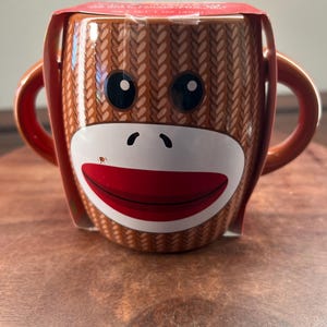May include: A brown ceramic mug with a sock monkey design. The mug features a white face, black eyes, and a red mouth. The mug has two orange handles and is packaged in a clear plastic wrap.