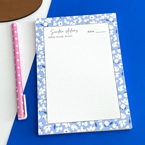May include: A floral-patterned notepad with the text "Sandra Adams" and "Bible Study Notes." The notepad has a light blue background with white flowers and a dotted interior. A pink pen with white polka dots is next to the notepad.