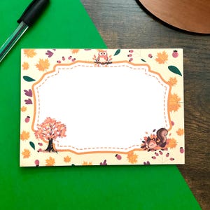 May include: A rectangular stationery card featuring a central white space framed by an orange border. The design includes autumn-themed illustrations of maple leaves, berries, a small owl perched on a branch, and a squirrel resting in foliage.