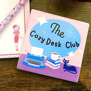May include: A pink square coaster with the text "The Cozy Desk Club" and illustrations of a typewriter, books, a mug, and a cat. A matching pen and notepad are visible in the background.