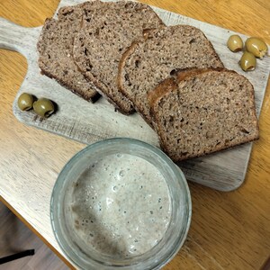 May include: Sliced brown bread on a wooden cutting board with green olives. A glass jar with a light-colored, bubbly mixture is in the foreground. The bread slices are thick and have a textured appearance, suggesting a rustic style.