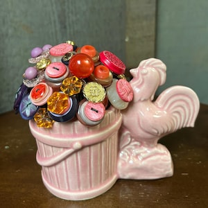 May include: A pink ceramic rooster figurine holding a pink ceramic bucket filled with colorful buttons. The buttons are a variety of shapes, sizes, and materials, including plastic, glass, and metal.