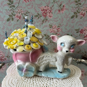 May include: A white ceramic lamb pulling a pink and blue wagon filled with yellow and white buttons. The wagon is decorated with a blue and white flower.