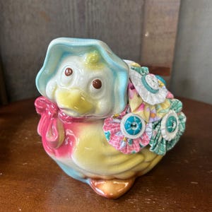 May include: A ceramic duck figurine with a blue bonnet and a yellow beak. The duck is decorated with colorful fabric flowers and buttons.