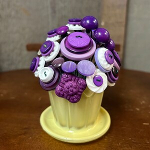 May include: A decorative button bouquet in a yellow ceramic pot. The arrangement features various purple and white buttons, creating a floral design. The pot sits on a matching saucer, and the background is a blurred wooden surface.