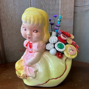 May include: A ceramic planter shaped like a girl with blonde hair, wearing a pink dress and a red bow. The planter is filled with colorful buttons and beads, creating a whimsical flower arrangement.