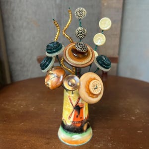 May include: A decorative button bouquet in a painted ceramic vase. The vase features a windmill scene. The bouquet includes various buttons in shades of green, brown, and cream, with gold wire stems. The arrangement is set on a wooden surface.