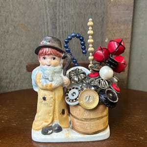 May include: A ceramic figurine of a shepherd holding a lamb, standing next to a small wooden barrel filled with buttons, beads, and bells. The figurine is painted in shades of brown, yellow, and white, and the barrel is painted in shades of brown and white.