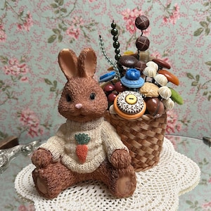 May include: A brown bunny figurine wearing a white sweater with a carrot design sits next to a wicker basket filled with colorful buttons and beads.