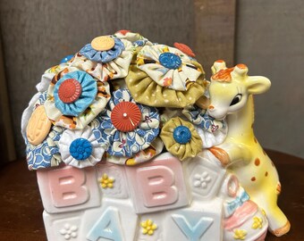 May include: A ceramic baby bank with a giraffe figurine and a stack of blocks spelling "BABY". The blocks are decorated with flowers and the giraffe is yellow with brown spots. The bank is topped with a cluster of colorful fabric rosettes.