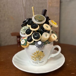 May include: A white teacup and saucer with a yellow bird and the words "Cliffy Falls" on the side. The teacup is filled with a bouquet of black, white, and green buttons.
