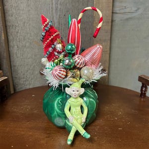 May include: A festive Christmas decoration featuring a green ceramic bag overflowing with ornaments, a red tree, and a candy cane. A seated elf figurine is in front of the bag. The ornaments include striped balls, green spheres, and a pink, ribbed teardrop.