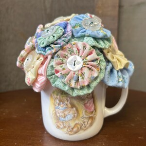 May include: A white ceramic mug with a Peter Rabbit design is decorated with a bouquet of fabric flowers. The flowers are made from colorful fabric scraps and have buttons in the center.