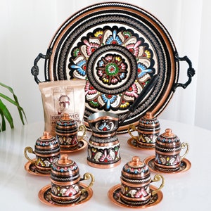 May include: A set of six hand-painted copper Turkish coffee cups and saucers with a matching copper coffee pot and a tray. The cups and saucers feature intricate floral designs in shades of pink, blue, green, and white on a black background. The coffee pot has a similar design and a black handle. The tray is round and has a black background with a floral design in shades of pink, blue, green, and white.