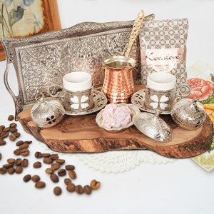 May include: A silver tray with an ornate design holds two small white coffee cups with silver saucers, a copper coffee pot, a silver sugar bowl, and a bag of Turkish coffee labeled "Kovanciye Turkish Coffee".