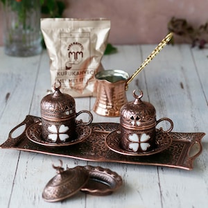 May include: A copper Turkish coffee set with two cups, saucers, and lids, a copper coffee pot with a gold handle, and a tray with intricate designs. The cups have white clover designs.