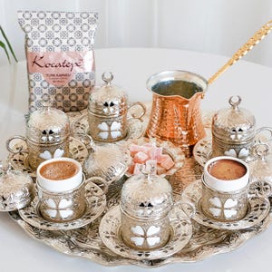 Może przedstawiać: Srebrna taca do serwowania z ozdobnym wzorem kwiatowym zawiera zestaw sześciu tureckich filiżanek do kawy i spodków. Każda filiżanka i spodek zdobione są wzorem koniczyny i białym wnętrzem. Na tacy znajduje się miedziana dzbanek do kawy ze złotą rączką oraz mała miseczka z tureckimi przysmakami. W tle widoczne jest opakowanie kawy "Kocatepe Turk Kahvesi".