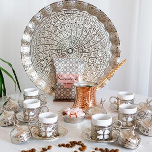 May include: A silver Turkish coffee set with a large, ornate tray, coffee cups, saucers, and lids. A copper coffee pot and a bag of coffee are also included. The set is arranged on a white surface, with coffee beans scattered around.