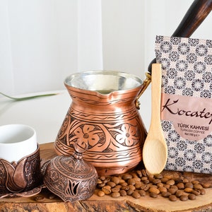 May include: A traditional Turkish coffee set with a copper cezve, a small copper cup and saucer, a wooden spoon, and a bag of Turkish coffee. The cezve has an intricate floral design and the cup and saucer have a similar design. The coffee bag is labeled "Kocatepe" and "Turk Kahvesi Turkish Coffee".