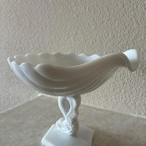 May include: A white milk glass compote dish featuring a scalloped rim and a bowl shaped like a shell. The base is hexagonal and supports a stem designed as a stylized fish or dolphin figure with textured scales, finished in a smooth opaque white.
