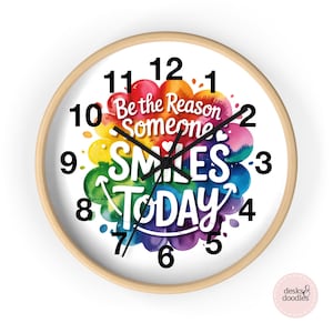 May include: A round wall clock with a light wood frame. The clock face is white with black numbers and hands. The center features a colorful watercolor design with the words "Be the Reason Someone Smiles Today."