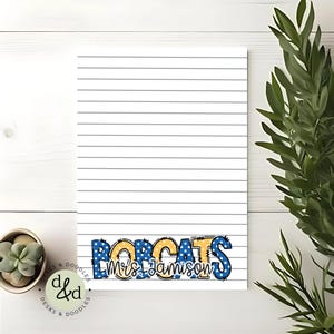 May include: Lined notepad with the text "BOBCATS Mrs. Jamison" in blue, yellow, and white. The notepad has a white background with gray lines. A small succulent plant and green leafy plant are in the background.