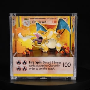 May include: A 3D pop-up card of a Charizard Pokemon card. The card is in a clear acrylic cube. The card features a yellow and orange Charizard with blue wings and a red tongue. The card text reads "Fire Spin Discard 2 Energy cards attached to Chariard in order to use this attack. 100"