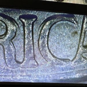 May include: Close-up of the word "RICA" in capital letters, likely from a coin or similar item. The letters are raised and have a textured surface, with a metallic appearance. The image is slightly out of focus.