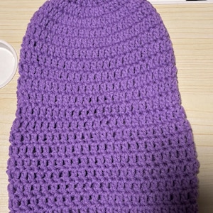 May include: A handmade, purple crochet beanie hat. The hat is made with a tight stitch pattern, giving it a textured appearance. The beanie is a solid color and appears to be made of soft yarn. The hat is designed to fit snugly on the head.