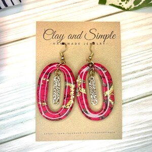 May include: A pair of red and gold oval-shaped earrings with a gold bar pendant hanging from each. The earrings are handmade and are displayed on a card that reads "Clay and Simple - Handmade Jewellery - https://www.facebook.com/ClayandSimple"
