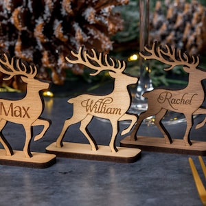 May include: Four wooden reindeer place card holders with the names Ashley, Max, William, and Rachel engraved on them. The reindeer are standing on a dark grey surface.
