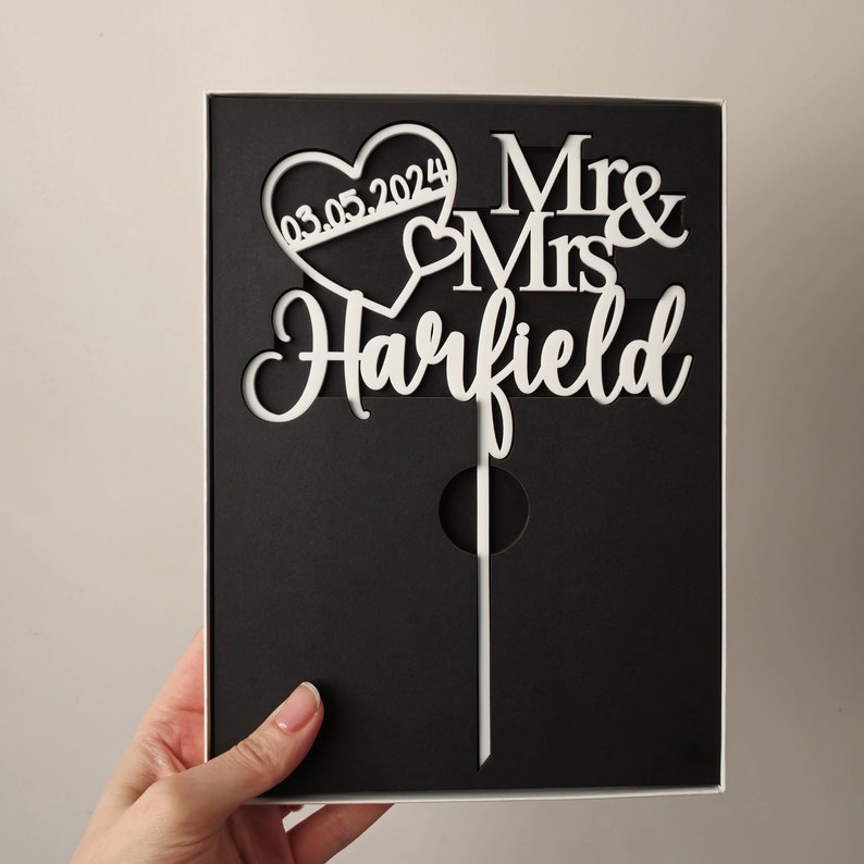May include: White cake topper with a heart design and the text "Mr & Mrs Harfield" and the date "03.05.2024".