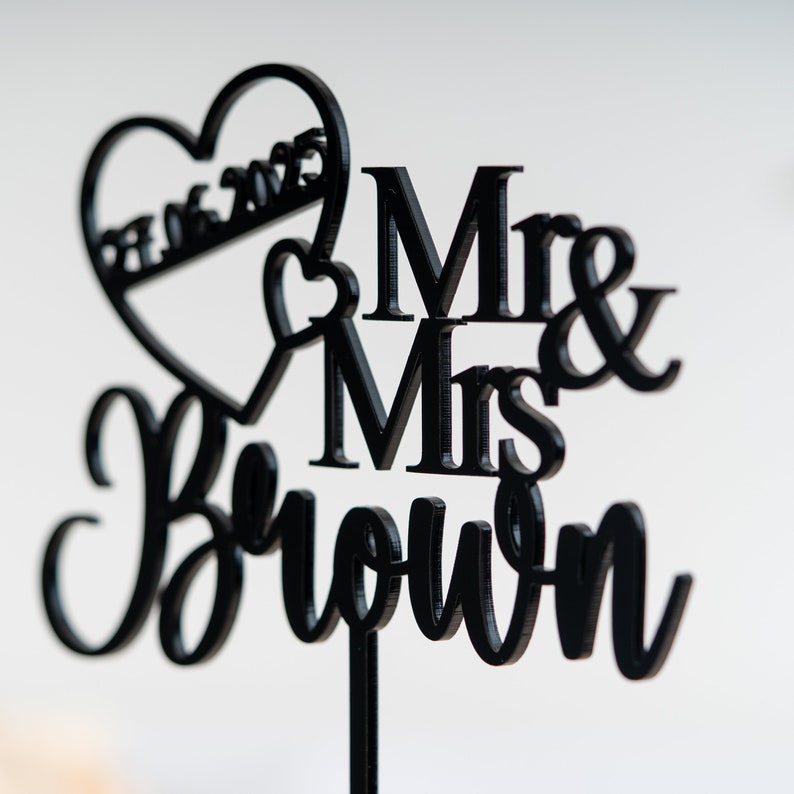 May include: Black acrylic cake topper with the words "Mr & Mrs Brown" and a heart with the date "07.16.2023" inscribed.