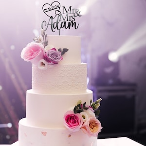 May include: A white wedding cake with four tiers decorated with pink and purple flowers. The top tier has a black cake topper that says "Mr & Mrs Adam" with a heart that says "15.10.2024".