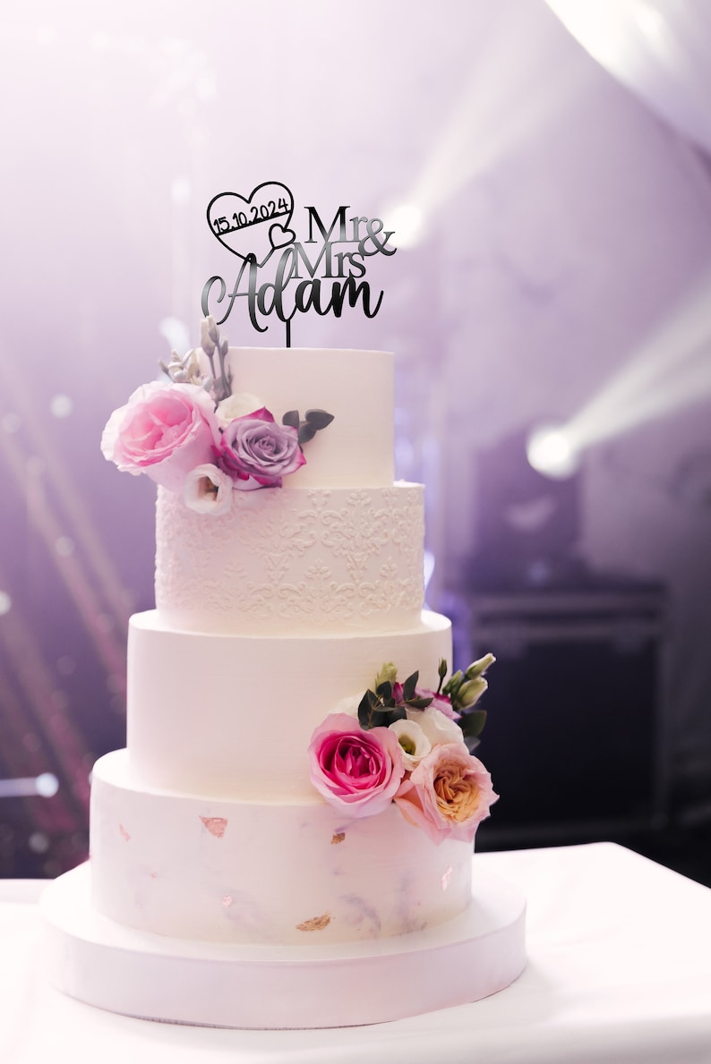 May include: A white wedding cake with four tiers decorated with pink and purple flowers. The top tier has a black cake topper that says "Mr & Mrs Adam" with a heart that says "15.10.2024".