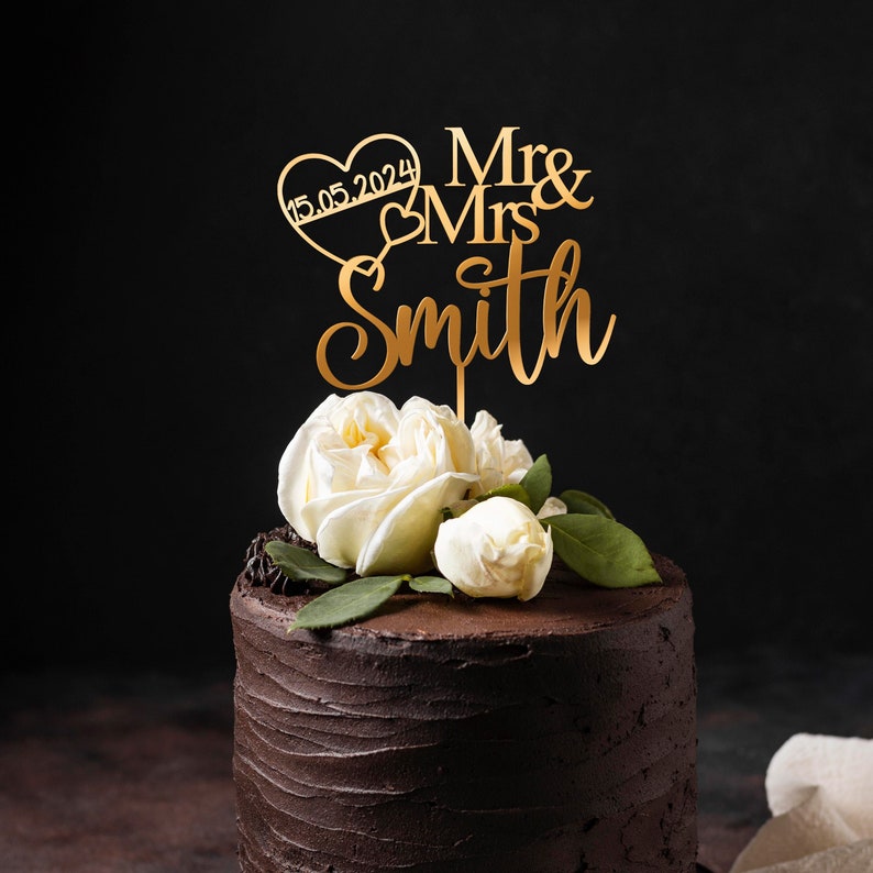 May include: Gold wedding cake topper with the text "Mr & Mrs Smith" and a heart with the date "15.05.2024". The topper is on a chocolate cake decorated with white roses.