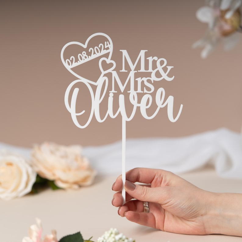 May include: White cake topper with a heart and the text "Mr & Mrs Oliver" and the date "02.08.2024".