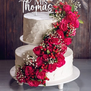 May include: Two-tiered white wedding cake decorated with a cascade of pink roses and white baby's breath. The cake topper is a white wooden sign that reads "Mr & Mrs Thomas" with a heart and the date "15.07.2024".
