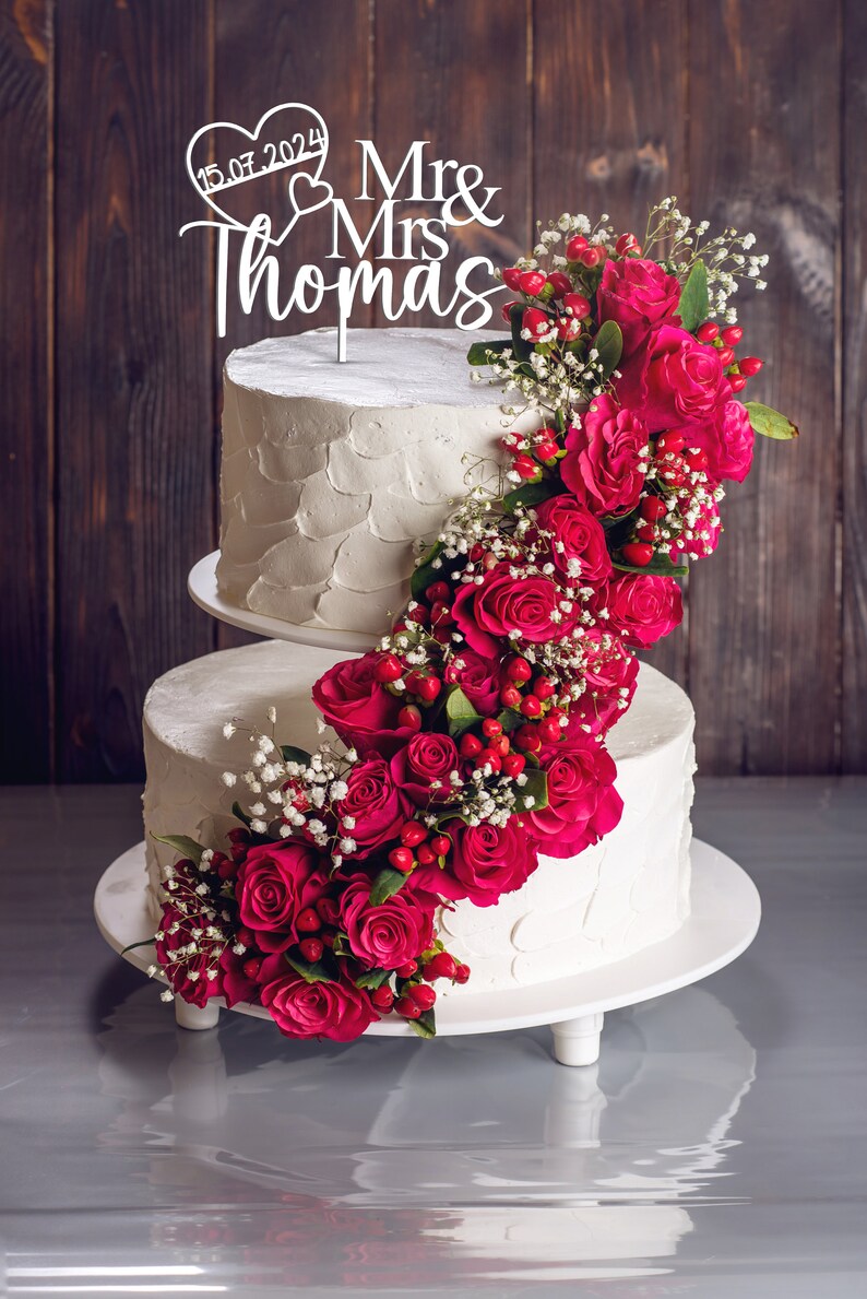 May include: Two-tiered white wedding cake decorated with a cascade of pink roses and white baby's breath. The cake topper is a white wooden sign that reads "Mr & Mrs Thomas" with a heart and the date "15.07.2024".