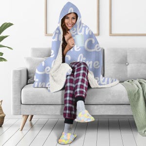 May include: A light blue hooded blanket with the word "celine" repeated in white text. The blanket is draped over a person sitting on a gray couch.