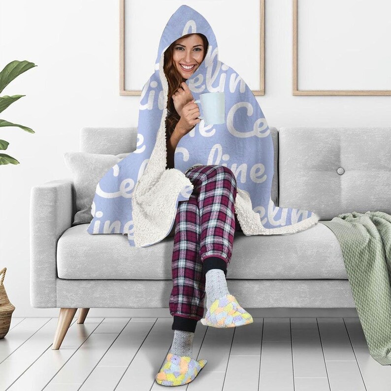 May include: A light blue hooded blanket with the word "celine" repeated in white text. The blanket is draped over a person sitting on a gray couch.