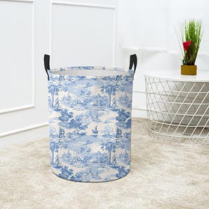 Pode incluir: Uma cesta de roupa azul e branca com alças pretas. A cesta é cilíndrica e tem um design de toile com fundo branco e figuras e árvores azuis.
