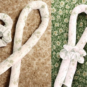 May include: White fabric hair curlers and a matching scrunchie, all adorned with a delicate floral pattern in shades of green. The curlers are long and cylindrical, designed for creating curls. The scrunchie is round and gathered.
