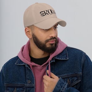 May include: A beige baseball cap with black embroidered text that reads "ERODA" on the front. The person wearing the cap is wearing a denim jacket over a pink hoodie.