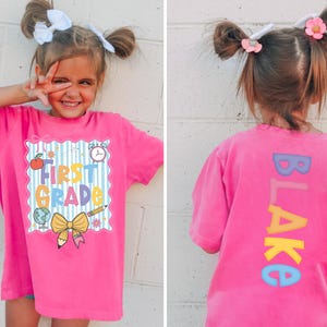 May include: A bright pink t-shirt with the words "FIRST GRADE" in a colorful design. The back of the shirt has the name "BLAKE" in vertical, colorful letters. The girl is smiling and wearing white bows in her hair.