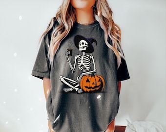 Può includere: Una maglietta grigia con una grafica di uno scheletro che indossa un cappello da strega e tiene una tazza di caffè. Lo scheletro è seduto su una zucca di Halloween.