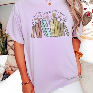 K&ouml;nnte beinhalten: Helllila T-Shirt mit dem Text "it's a good day to read a book" &uuml;ber einer Grafik von B&uuml;chern und Blumen. Das Shirt hat eine lockere Passform und einen bequemen Stil.
